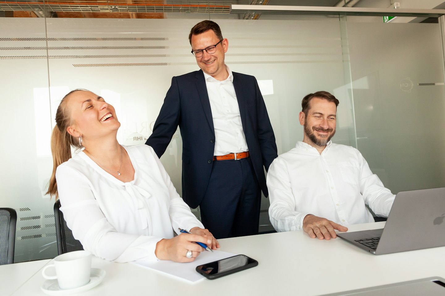 Tres profesionales sonriendo colaboran alrededor de una mesa en una oficina moderna.